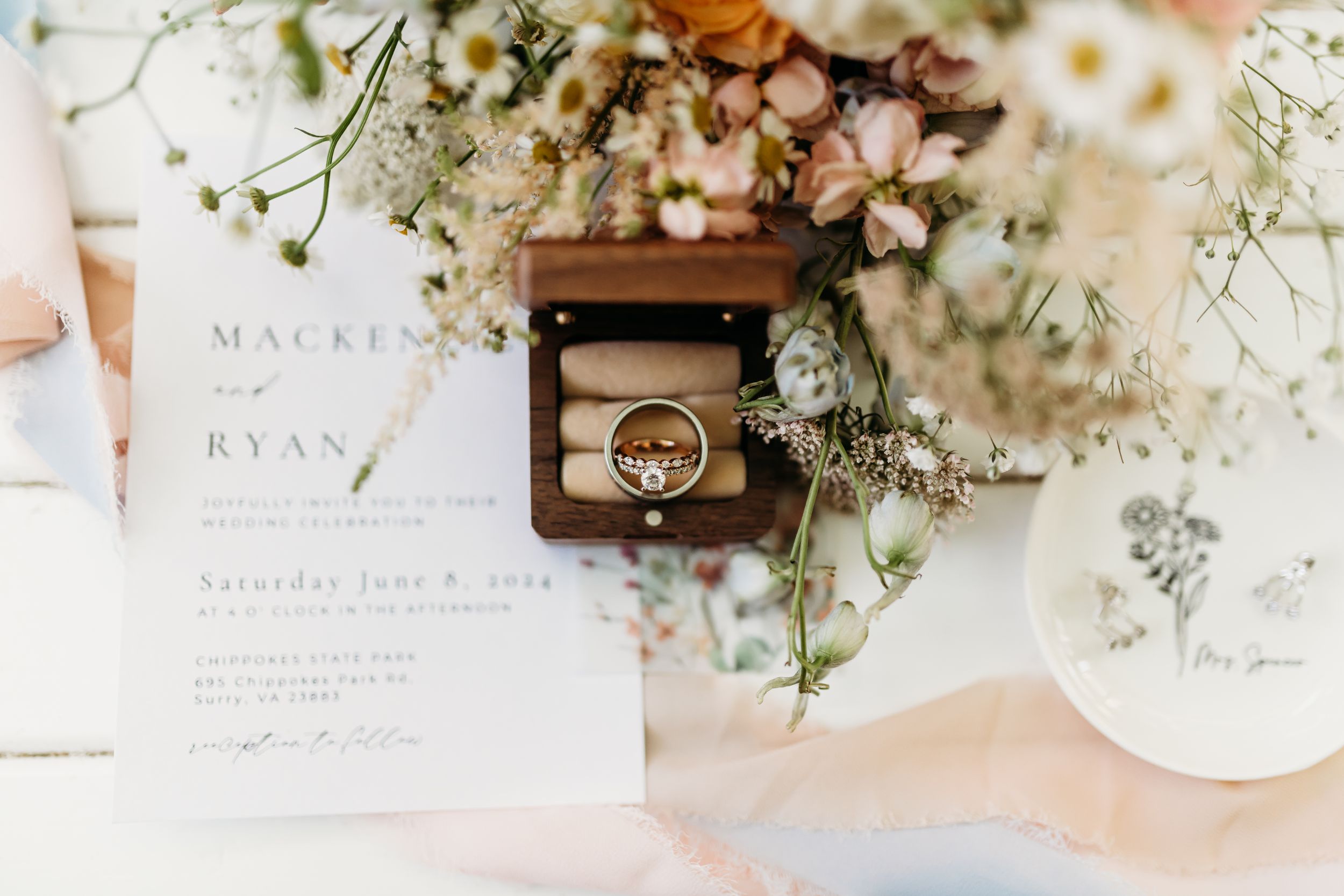 Wedding invitation and rings styled with delicate flowers and greenery on light background.