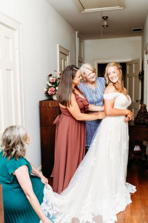 Wedding party helps bride with dress preparations in indoor hallway setting.