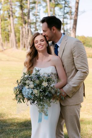 A couple shares a tender moment outdoors with a stunning blue and white wedding bouquet in a wooded setting.