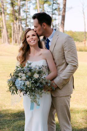 Newlywed couple shares romantic moments in a sun-drenched pine forest setting with elegant floral bouquet details.