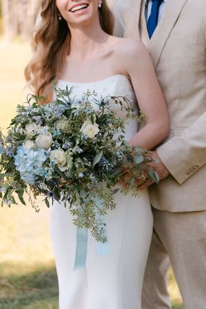 Close-up of an elegant bridal bouquet featuring blue hydrangeas, white roses and greenery against a white wedding dress.