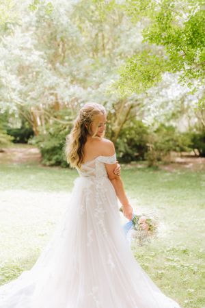 Elegant bride in off-shoulder gown holding bouquet while looking over shoulder in garden setting.