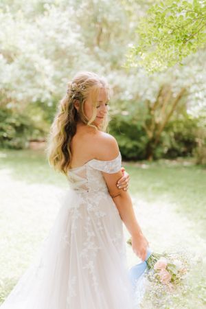 Bride in lace off-shoulder gown holds bouquet in sunlit garden setting.