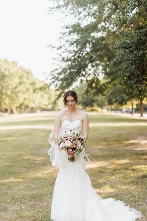 Bride in strapless mermaid gown holding pink floral bouquet, standing on green lawn surrounded by trees.