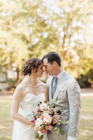 Bride and groom share a romantic moment outdoors, she holds a pink bouquet, he wears a gray suit with blue tie.