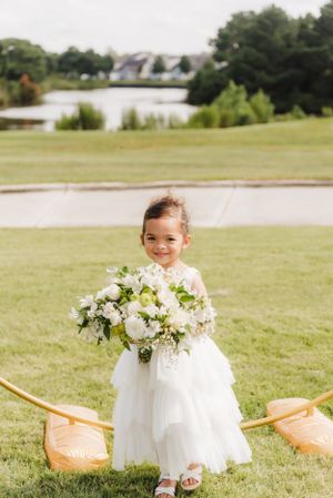 Young flower girl in white dress holding a beautiful bouquet of white flowers on a grassy lawn.