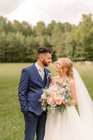 A romantic wedding couple shares an intimate moment in a sunlit field with the bride holding a peach and white bouquet.