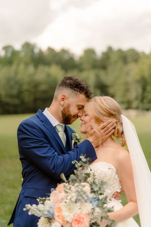 Romantic wedding couple shares intimate moments in a lush outdoor setting with peach and blue floral bouquet.