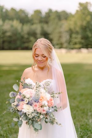 A bridal bouquet features soft peach roses, white flowers and eucalyptus greenery against a dreamy outdoor background.