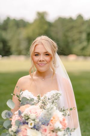 A bride holds a beautiful bouquet of pink and blue flowers against a soft green outdoor backdrop.