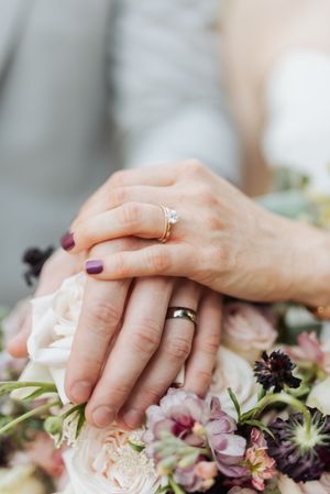 Bride and groom hands with wedding rings resting on a bouquet of pink and purple flowers.