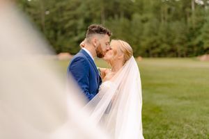 A romantic wedding moment captured outdoors with a flowing white veil and blue suit against a green natural backdrop.
