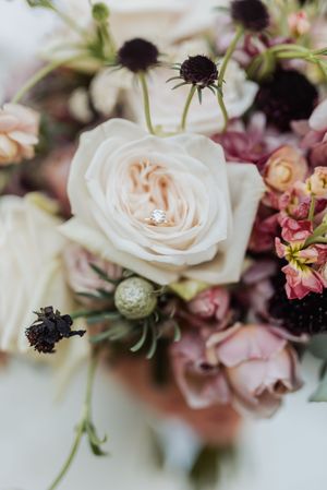 A diamond engagement ring nestled in a lush bridal bouquet of cream roses, dusty pink blooms, and dark thistle accents.