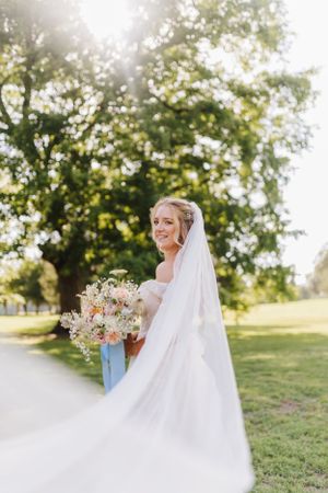 Beautiful bride in flowing white dress and veil holding bouquet outdoors in sunlit garden setting.