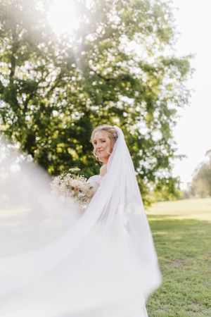 Side profile of bride holding bouquet with flowing veil in sunlit outdoor garden.