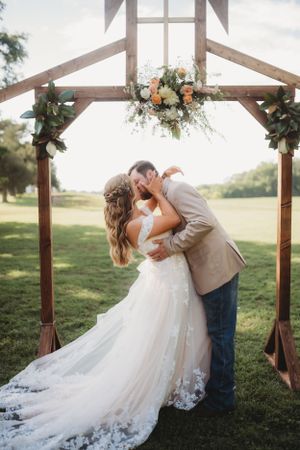 Romantic wedding kiss under rustic wooden arch decorated with peach and white floral arrangement.