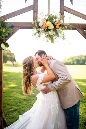 Passionate wedding kiss under wooden arch adorned with peach and white florals.
