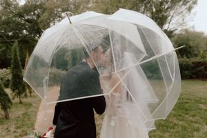 A romantic couple shares a kiss under a clear umbrella in a rainy outdoor setting with lush greenery.