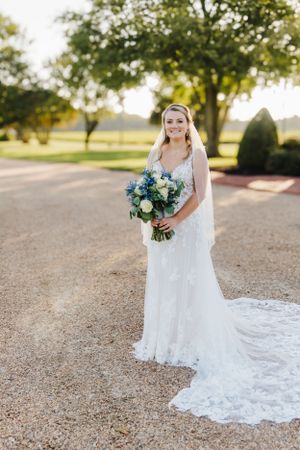 A bride in a flowing lace wedding dress holds a blue and white bouquet on a gravel path.
