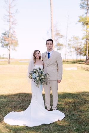 A wedding couple poses together on a grassy hill with tall pine trees and golden sunlight in the background.