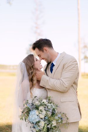 A romantic wedding couple embraces outdoors while holding a blue and white floral bouquet against a sunny autumn backdrop.
