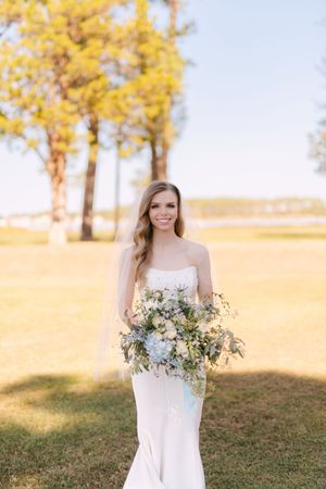 A bride in a white gown holds a beautiful blue and white bouquet against a backdrop of golden autumn trees.
