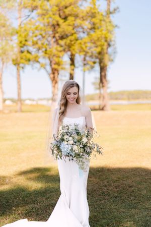 A bride in a flowing white dress stands in a field during golden hour with autumn foliage in the background.