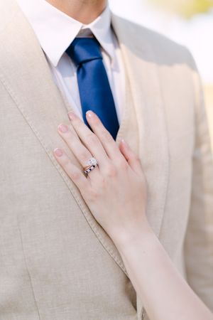 Close-up detail of a light beige suit jacket with navy tie and a diamond ring on a hand.