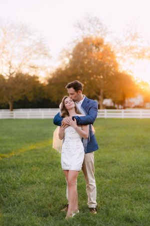 Couple dancing and embracing in a grassy field at sunset with white fencing in the background.