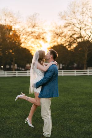Wedding couple shares romantic sunset kisses in a field with white fence in background.