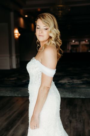 Person in off-shoulder lace wedding dress stands in elegant dark venue.