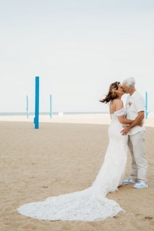 Couple embraces on beach with flowing white wedding dress and blue posts in background.