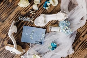 Wedding details flatlay with blue flowers, veil, and rings on rustic wooden surface.
