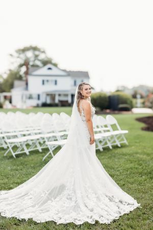 Bride in lace gown poses outdoors with white ceremony chairs and house in background.