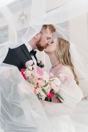A romantic wedding photograph with flowing veil and pink bouquet flowers captured in soft natural light.