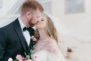 A romantic couple shares a tender kiss during a wedding celebration, with pink flowers and soft natural lighting.
