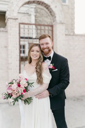 A couple embraces on their wedding day, the bride holding a pink floral bouquet against a stone archway background.