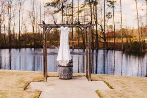 A white wedding dress hangs on a wooden arch overlooking a serene lake at sunset.