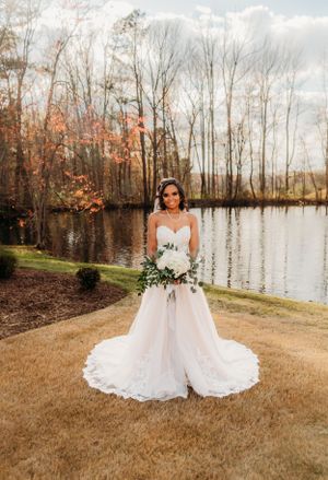 Elegant bride stands alone by waterfront in flowing lace gown with trailing bouquet.