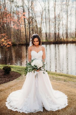 Bride in lace wedding gown poses by lake during fall with white bouquet.