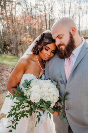 Newlyweds share tender moment while holding elegant white floral arrangement.