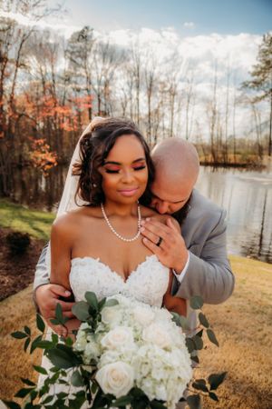 Romantic wedding couple embraces by lakeside during autumn with white floral bouquet.
