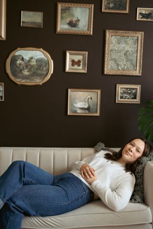 A person lounges in white sweater and blue jeans on a brown sofa against a wall decorated with framed artwork.