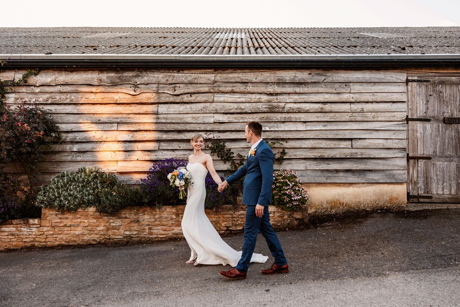 A bride and groom dance together at sunset in front of a rustic wooden barn wall.