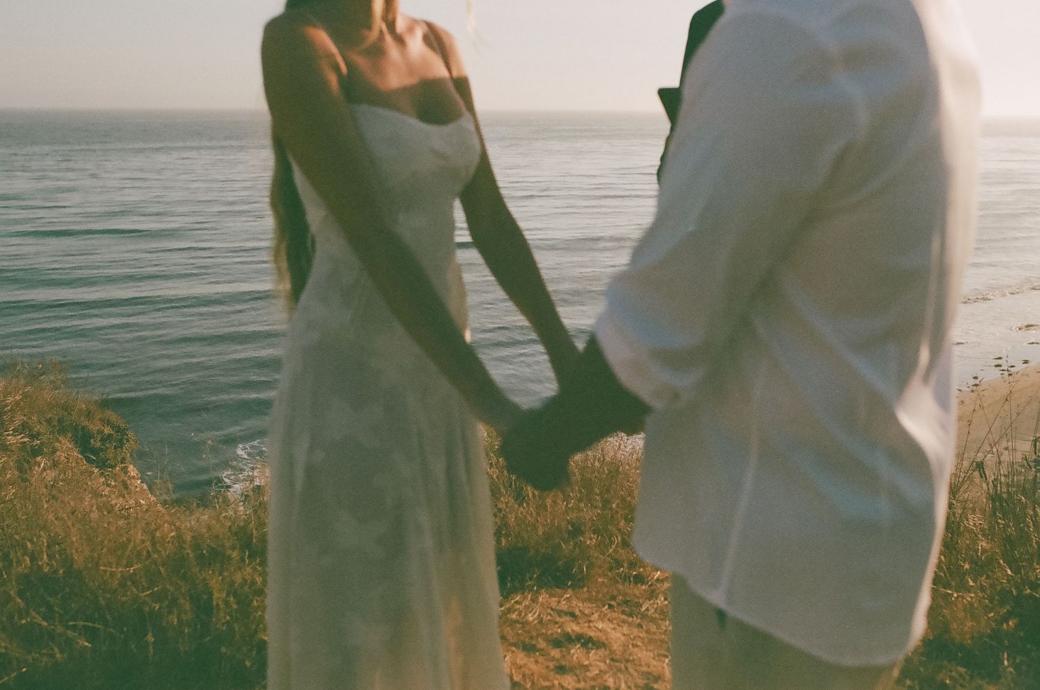 A couple walks hand in hand along a serene beach at sunset with waves rolling in the background.