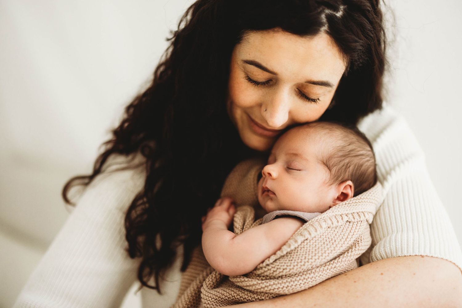 A tender moment captured as mother and newborn baby snuggle together in soft white and beige tones.