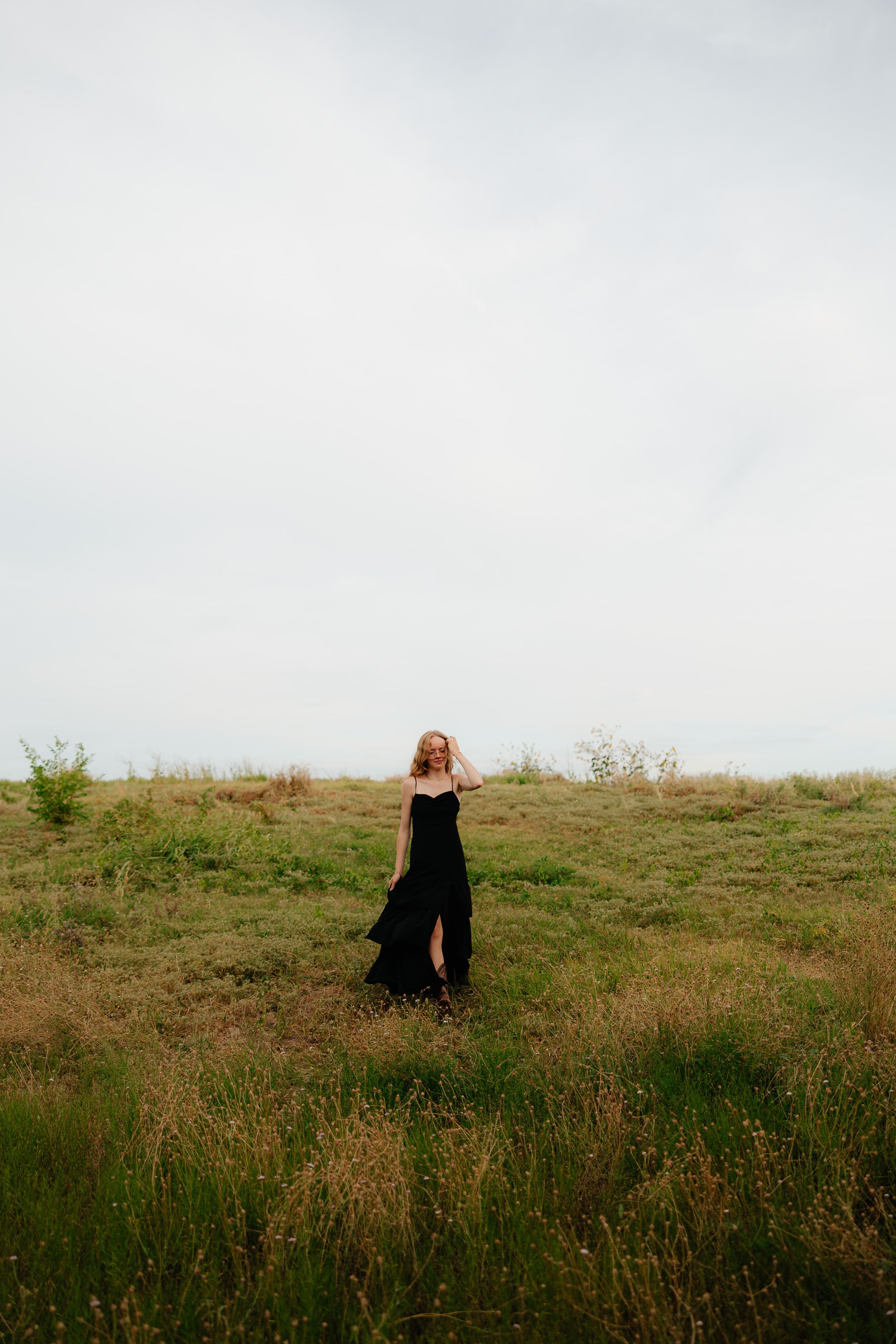 Person in black dress twirls in grassy field against cloudy sky.