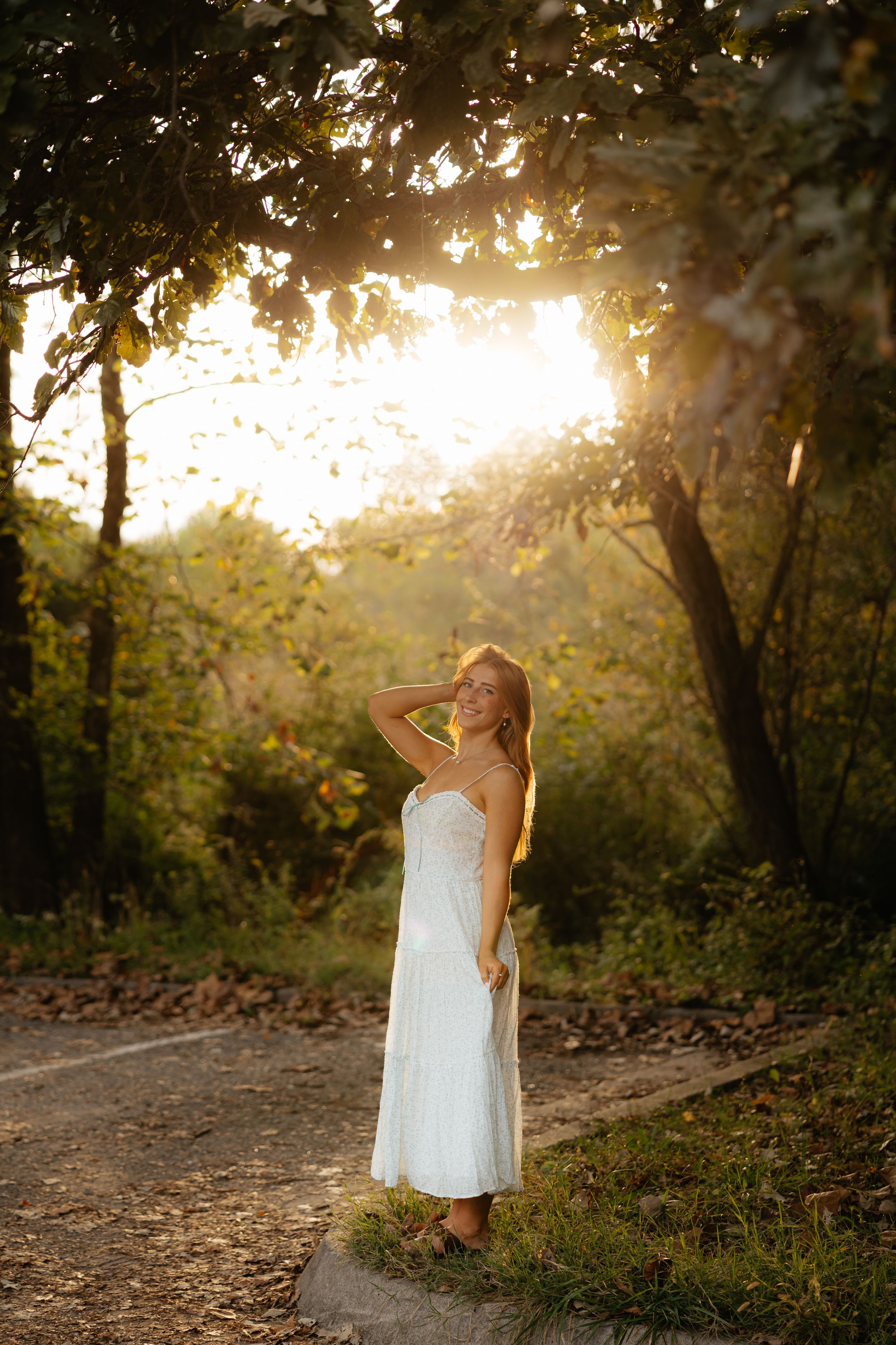 Figure in white dress poses on nature trail during sunset with sunlight streaming through trees.