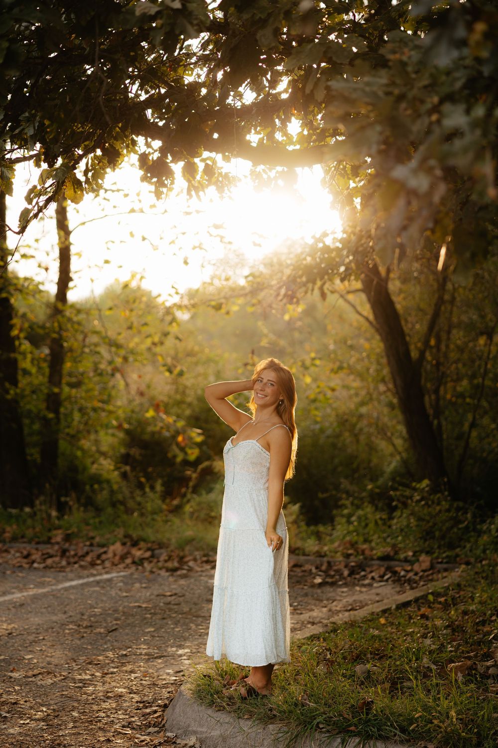Figure in white dress poses on nature trail during sunset with sunlight streaming through trees.