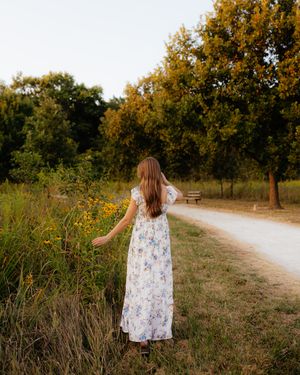 Woman in floral dress stands on dirt path surrounded by autumn trees and wildflowers at golden hour.